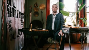 Sam Wenc in teal henley stands in home studio with pedal steel guitar, plants, hanging metalwork, and text banner, warm lighting.