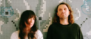 Two members of Special Friend against mosaic tile wall in green, gray, and white tones. One wears white shirt, one dark sweater.